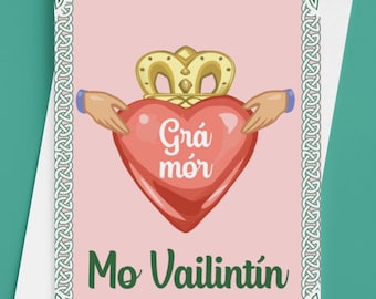 May include: A greeting card with a pink background features a red heart topped with a gold crown, held by two hands. The heart has the words "Grá mór" in white. Below the heart, the words "Mo Vailintín" are written in green. The card is framed with a green Celtic knot design.