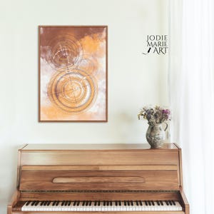May include: Abstract art print with a brown and orange background and white geometric shapes. The print is framed in a light brown wood frame and is hanging on a white wall above a wooden piano.