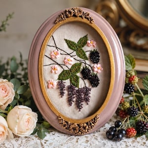 May include: An oval embroidery hoop frame in dusty rose with gold accents, featuring a needlework design on beige fabric. The embroidery depicts green leaves, pink flowers, and clusters of black and purple berries with intricate beadwork details.