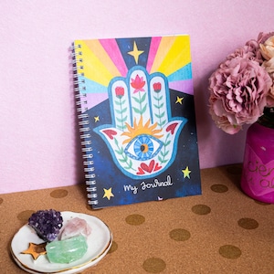 May include: A spiral bound journal featuring a colourful illustration of a hamsa hand with an eye in the centre. The background is a dark blue with stars and a rainbow. The text "My Journal" is printed on the cover.