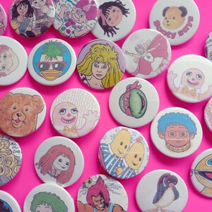 May include: A collection of colorful round buttons featuring various cartoon characters, including a bear, a penguin, a turtle, and a duck. The buttons are arranged on a pink background.