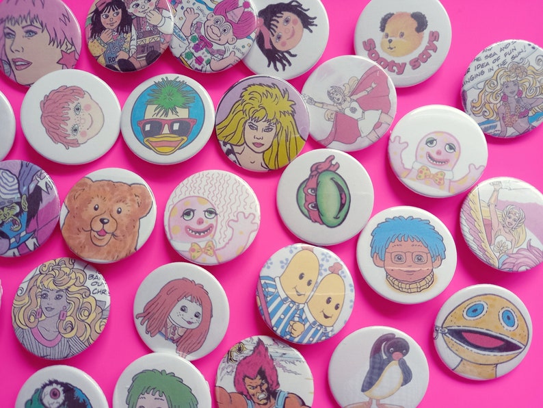 May include: A collection of colorful round buttons featuring various cartoon characters, including a bear, a penguin, a turtle, and a duck. The buttons are arranged on a pink background.