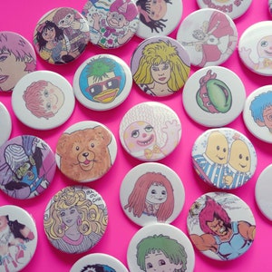 May include: A collection of colorful round buttons featuring various cartoon characters, including a bear, a penguin, a duck, and a banana. The buttons are arranged on a pink background.