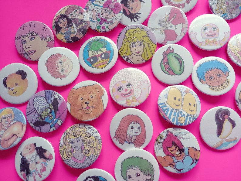May include: A collection of colorful round buttons featuring various cartoon characters, including a bear, a penguin, a duck, and a banana. The buttons are arranged on a pink background.