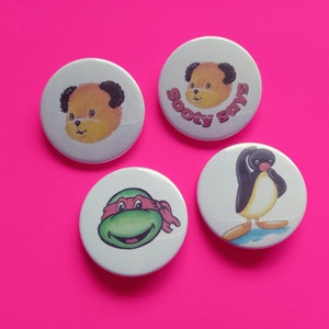 May include: Four round white button badges with different cartoon characters. The first badge features a yellow bear with black ears and the text "Sooty Says". The second badge features a green turtle with a red bandana. The third badge features a yellow bear with black ears. The fourth badge features a black and white penguin.