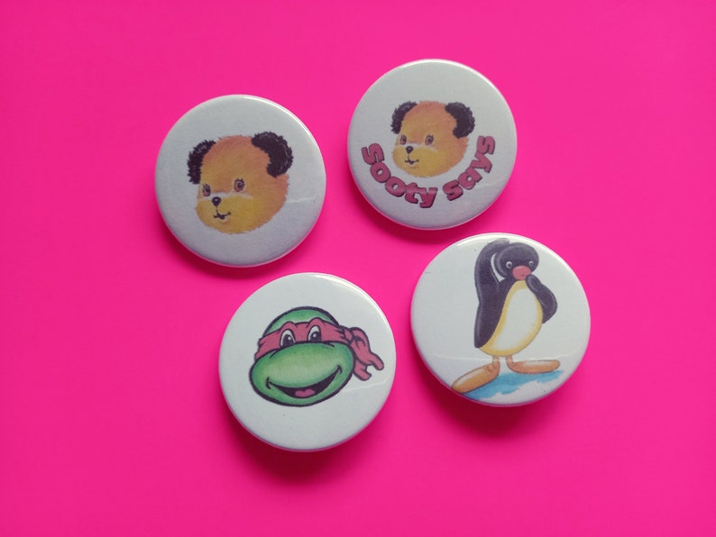 May include: Four round white button badges with different cartoon characters. The first badge features a yellow bear with black ears and the text "Sooty Says". The second badge features a green turtle with a red bandana. The third badge features a yellow bear with black ears. The fourth badge features a black and white penguin.