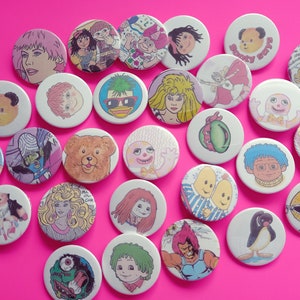 May include: A collection of 25 colorful round pinback buttons featuring various cartoon characters and illustrations. The buttons are approximately 1 inch in diameter and have a white background.