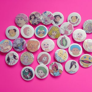 May include: A collection of colorful round buttons featuring cartoon characters and illustrations. The buttons are made of metal and have a shiny finish.