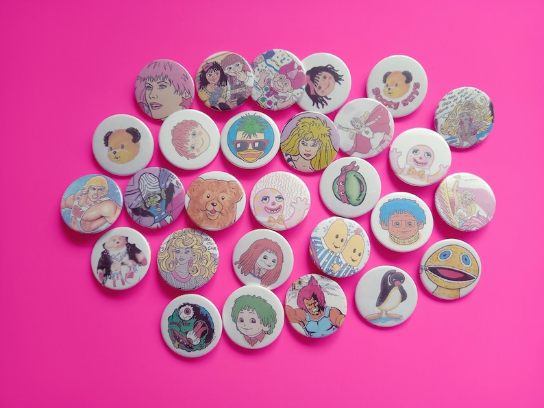 May include: A collection of colorful round buttons featuring cartoon characters and illustrations. The buttons are made of metal and have a shiny finish.