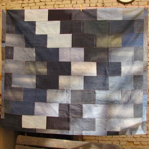 May include: A large patchwork quilt made from denim fabric in various shades of blue. The quilt is hanging on a clothesline against a brick wall.