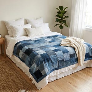 Op de afbeelding: Een patchwork sprei gemaakt van verschillende tinten blauwe denim stoffen vierkanten bedekt een bed met wit beddengoed. Een crèmekleurige gebreide deken ligt over de sprei. Het bed staat in een kamer met een lichte houten vloer en witte muren.