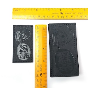 Puede incluir: Dos parches de cuero negro con texto blanco y un gráfico de calavera. El texto en los parches dice "Cheap Monday".