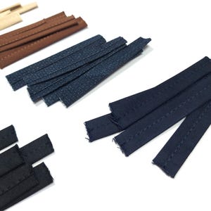 May include: Assortment of fabric strips in various colors, including beige, brown, navy, and black. The strips are rectangular and appear to be pre-cut for sewing or crafting projects. The image is shot on a white background.