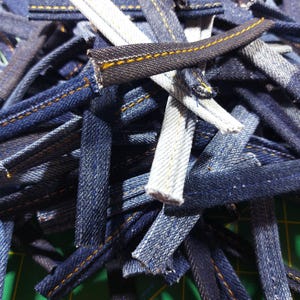 May include: A pile of denim fabric strips with yellow stitching. The strips are various shades of blue and some are white.