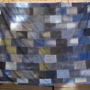 Può includere: Una trapunta patchwork in denim, di forma rettangolare, con un motivo a mattoni. La trapunta è composta da quadrati di denim di varie tonalità di blu e grigio. La trapunta è appesa, mostrando le sue dimensioni e la sua consistenza.