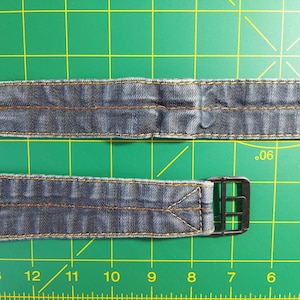 Puede incluir: Dos correas de reloj de mezclilla con una hebilla negra. Las correas están cosidas con hilo amarillo. Las correas están sobre una estera de corte verde con una regla.