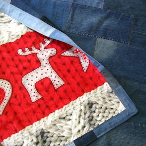 May include: A red and white fabric with a knitted pattern and a white reindeer with polka dots. The fabric is sewn to a blue denim fabric with a patchwork design.