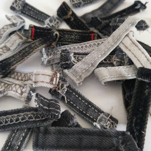 May include: A collection of assorted denim belt loops in various shades of black, gray, and white. Each strip features visible stitching along the edges, with some pieces showing frayed ends and one loop accented with a small red thread detail.