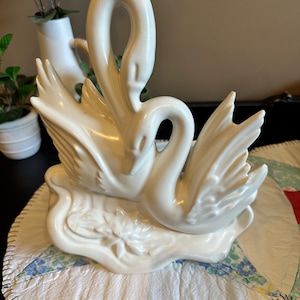 以下が含まれることがあります： 白磁の白鳥の置物。2羽の白鳥が絡み合い、翼を広げ、首でハート型を形成しています。置物はスイレンの葉の台座に置かれています。家の装飾用の装飾品。