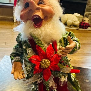 May include: A whimsical Christmas elf figurine with a green and gold patterned outfit, a red hat, and a large red poinsettia flower. The elf is smiling and has a white beard.