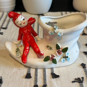 Könnte beinhalten: Ein Vintage-Keramik-Blumentopf in Form eines weißen Stiefels, verziert mit rosa und blauen Blumen und grünen Blättern. Eine rote Figur einer Person in einem roten Overall und Hut lehnt an dem Stiefel. Der Pflanztopf steht auf gemustertem Stoff.