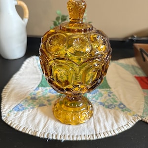 以下が含まれることがあります： 琥珀色のガラス製キャンディーディッシュ、蓋付き。星と花の模様が施された丸く質感のあるボディが特徴です。台座の上にあり、蓋にはお揃いのつまみが付いています。このディッシュはおそらくヴィンテージで、エレガンスを添えています。