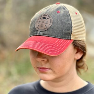 May include: A black and tan baseball cap with a red brim. The cap has a brown leather patch with the text "Nebraska" and a compass design with the letters "N" and "E" and the year "1867".