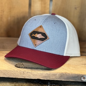 May include: A gray and white trucker hat with a burgundy brim. The hat features a brown leather patch with a fish silhouette in the center.