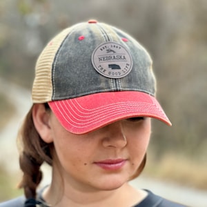 May include: A black and tan distressed baseball cap with a red brim. The cap has a brown leather patch with the text "Est. 1867 Nebraska The Good Life" in white.