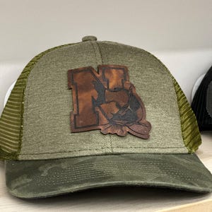 May include: A green and brown trucker hat with a camouflage brim. The hat features a brown leather patch with the letter "N" and a fish design. The mesh back is olive green.