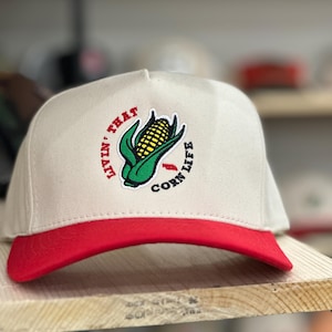 May include: A beige baseball cap with a red brim. The cap features an embroidered patch with a green corn cob and the text "Livin' That Corn Life".
