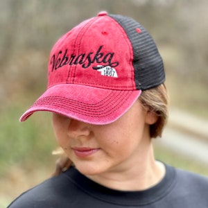 May include: A red and black baseball cap with a black embroidered logo that says "Nebraska Est. 1867".