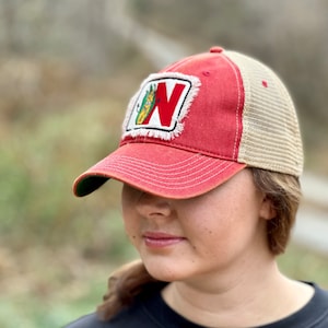 May include: Red and tan trucker hat with a patch featuring a white-bordered "N" and a corn graphic. The hat has a red brim and crown, with a tan mesh back. The hat is worn by a person.