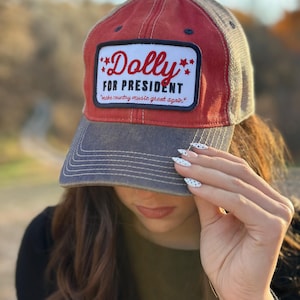 May include: A red and grey baseball cap featuring a rectangular patch on the front. The patch has a white background with the red text Dolly, the black text FOR PRESIDENT, and the smaller red text make country music great again with red stars.