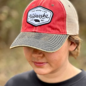 May include: A red and beige baseball cap with a black and white patch that says "The Good Life Nebraska Est. 1867".