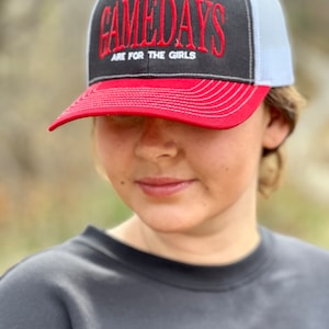 May include: A red, white, and gray baseball cap with a black mesh back. The front of the cap has the text "GAMEDAYS ARE FOR THE GIRLS" in red and white embroidery.