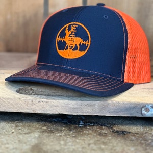 May include: A black and orange trucker hat with a deer silhouette inside a target graphic. The hat has a mesh back.