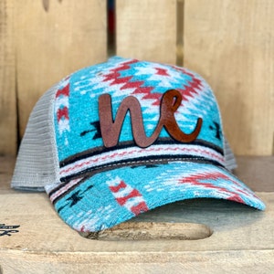 May include: A turquoise, red, and white patterned trucker hat with a brown leather "me" applique.