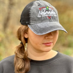 May include: A black and gray distressed baseball cap with a black mesh back. The cap has a white embroidered logo that says "Nebraska The Cornhusker State" with a red and green corn stalk graphic.