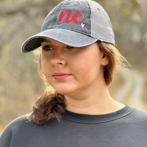 May include: A black and gray baseball cap with a red embroidered "me" and a small white heart. The cap has a mesh back.