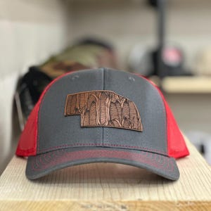 May include: A gray and red trucker hat with a brown leather patch of the state of Nebraska with corn stalks on it.