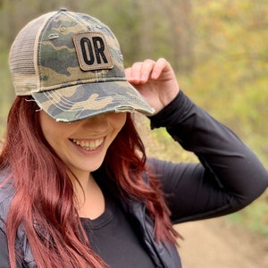 May include: A camouflage trucker hat with a distressed look and a brown leather patch that says "OR" in black letters.