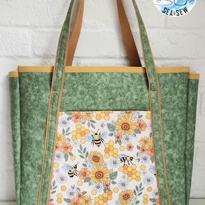 Peut inclure: Un sac fourre-tout avec un corps en tissu vert chiné, une bordure jaune moutarde et deux anses en tissu marron. Le panneau avant présente un fond blanc avec un motif répétitif de tournesols jaunes, de fleurs orange, de rayons de miel et d'abeilles.