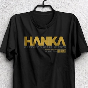 May include: Black unisex t-shirt with the gold text "HANKA PRECISION INSTRUMENTS" and Japanese text. The shirt is hanging on a black hanger. The bottom of the shirt has the text "UNISEX T-SHIRT".