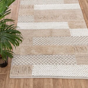 May include: A beige and white woven rug with a geometric pattern. The rug has a fringe border and is made of natural fibers.