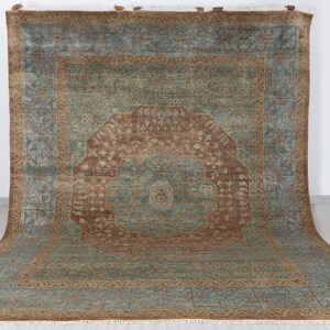 May include: A large, rectangular rug with a central medallion design. The rug features a teal and brown colour palette with intricate patterns and a fringed edge. The rug is displayed on a light-coloured floor.