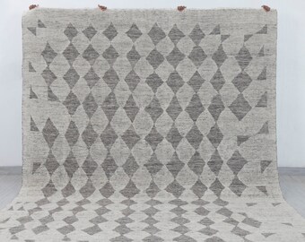 Pode incluir: Um tapete cinza claro com um padrão de losangos repetido em cinza mais escuro. O tapete tem uma borda com franjas e é exibido em um fundo branco. O tapete é feito de um material tecido.