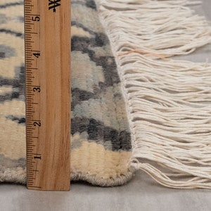 Puede incluir: Primer plano del borde de una alfombra de color crema con un patr&oacute;n geom&eacute;trico negro y gris. La alfombra tiene un borde de flecos y se mide con una regla de madera.
