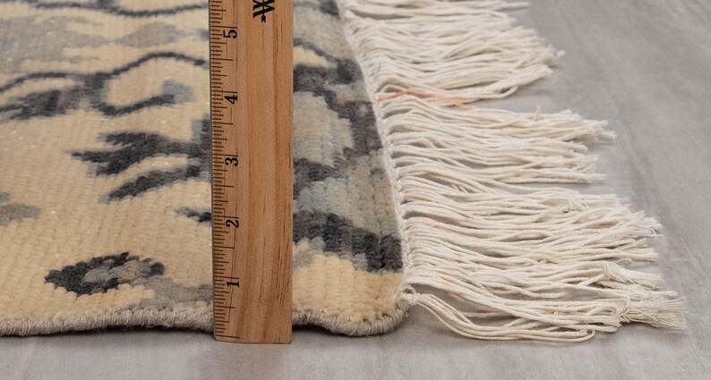 Puede incluir: Primer plano del borde de una alfombra de color crema con un patr&oacute;n geom&eacute;trico negro y gris. La alfombra tiene un borde de flecos y se mide con una regla de madera.