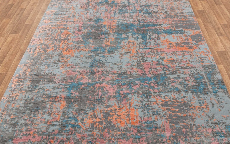 K&ouml;nnte beinhalten: Ein gro&szlig;er, rechteckiger Teppich mit einem abstrakten Design. Der Teppich hat eine graue Grundfarbe mit Farbspritzern in Blau, Orange und Rosa. Das Muster hat ein verwittertes, strukturiertes Aussehen, geeignet f&uuml;r ein Wohn- oder Schlafzimmer.